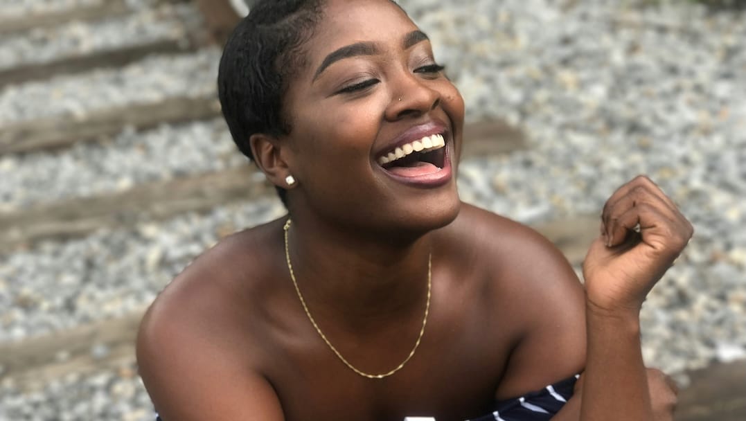 Joyful Black woman laughing with head tilted back, bare shoulders, gold chain necklace, outdoors near wooden steps and gravel backdrop.